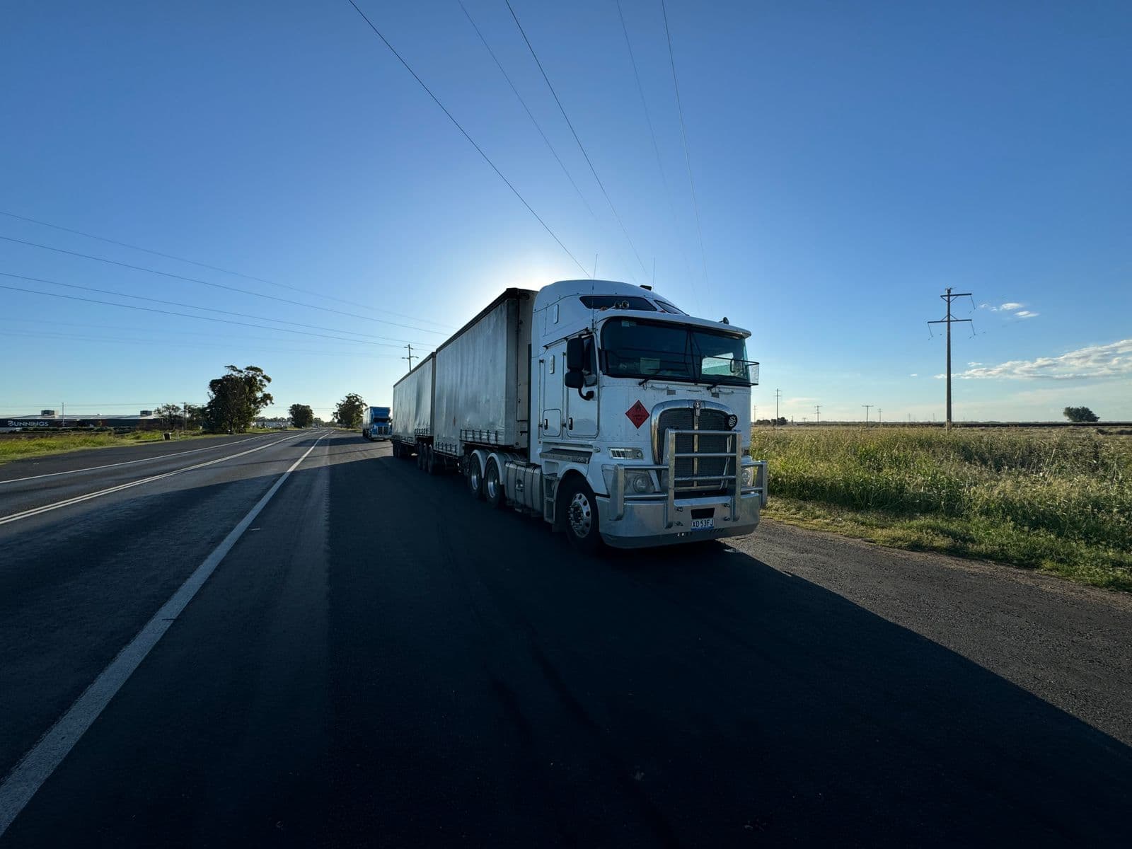A white prime mover pulling two trailers on a rural Australian highway under a clear blue sky